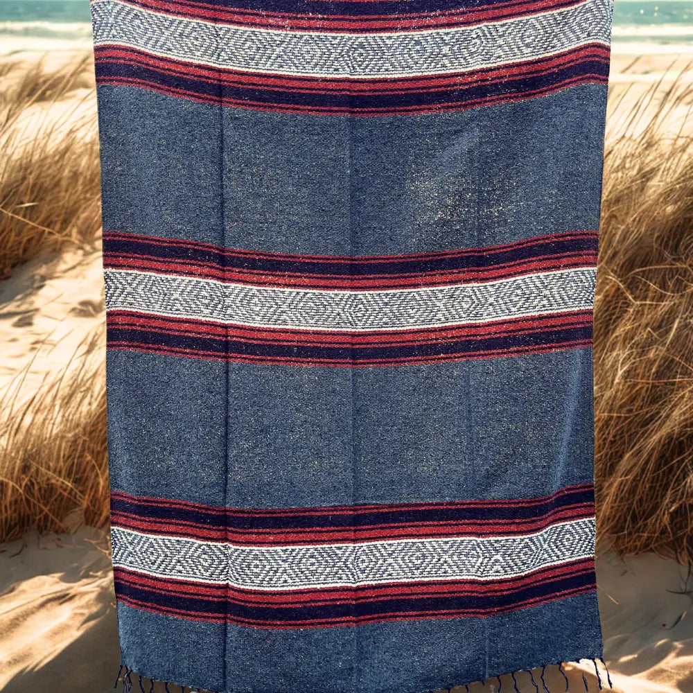 The Faire Authentic Mexican Blanket Ember/Grey, featuring geometric patterns, hangs vertically outdoors with sand, dry grass, and water in the background.