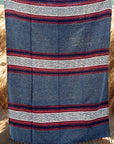 The Faire Authentic Mexican Blanket Ember/Grey, featuring geometric patterns, hangs vertically outdoors with sand, dry grass, and water in the background.