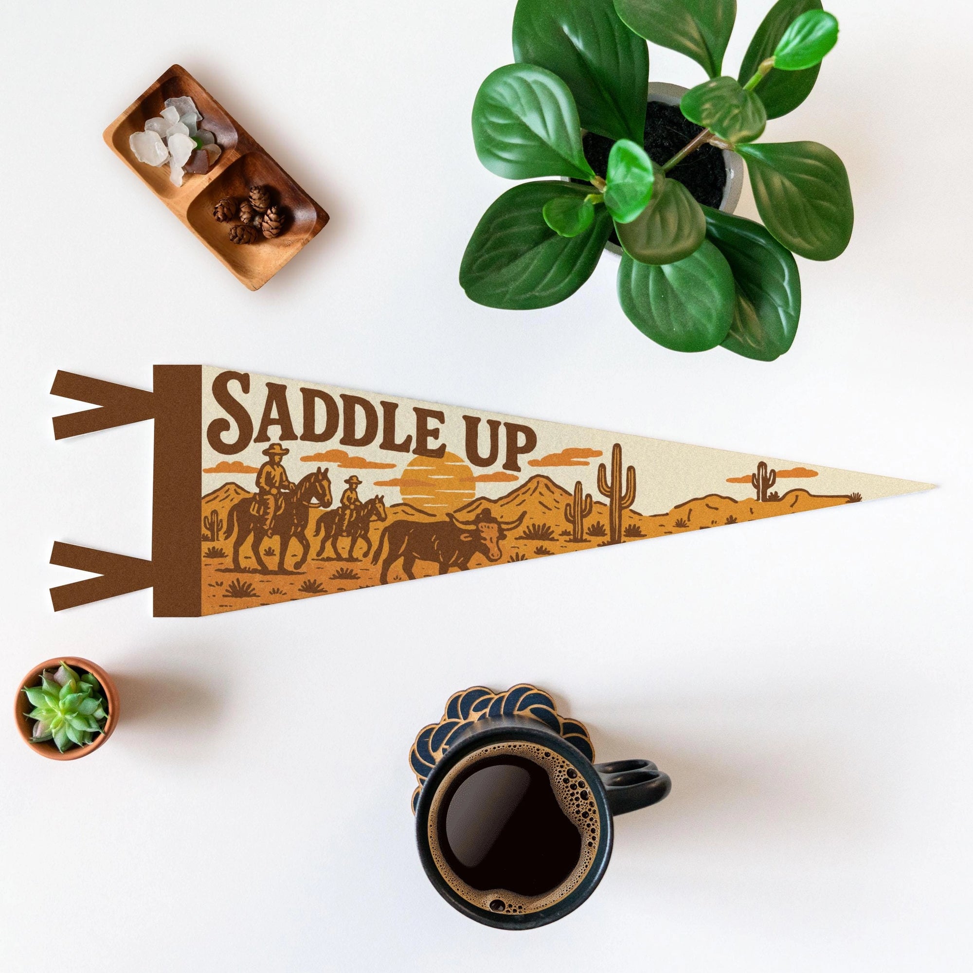 The "Saddle Up" Felt Pennant by Faire, featuring a cowboy scene, rests on a white surface with coffee, green plants, and crystals—ideal as western decor or a gift for horse lovers.