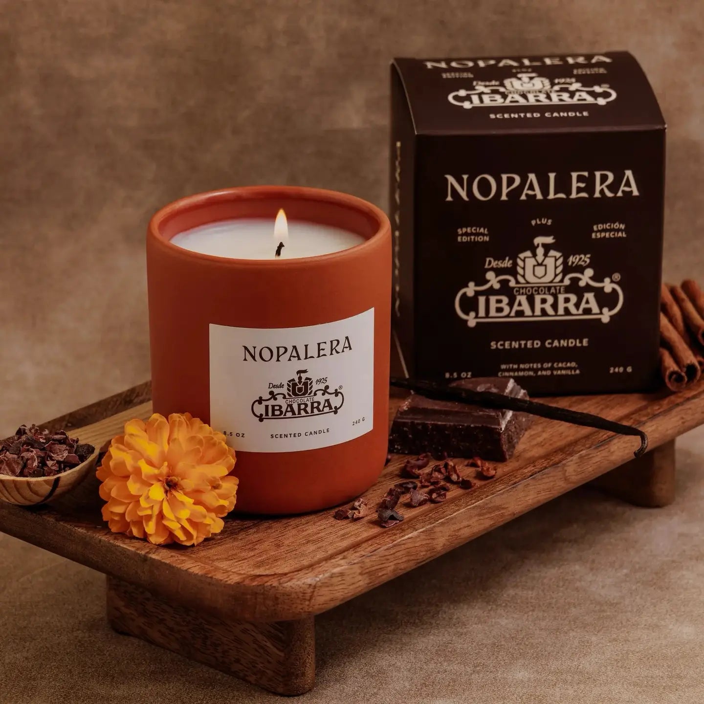 A Nopalera Chocolate Candle by Faire, in a brown glass jar, sits on a tray with marigold, cinnamon sticks, and chocolate pieces. The box highlights the “Chocolate Ibarra” scent inspired by Mexican heritage.