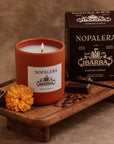 A Nopalera Chocolate Candle by Faire, in a brown glass jar, sits on a tray with marigold, cinnamon sticks, and chocolate pieces. The box highlights the “Chocolate Ibarra” scent inspired by Mexican heritage.