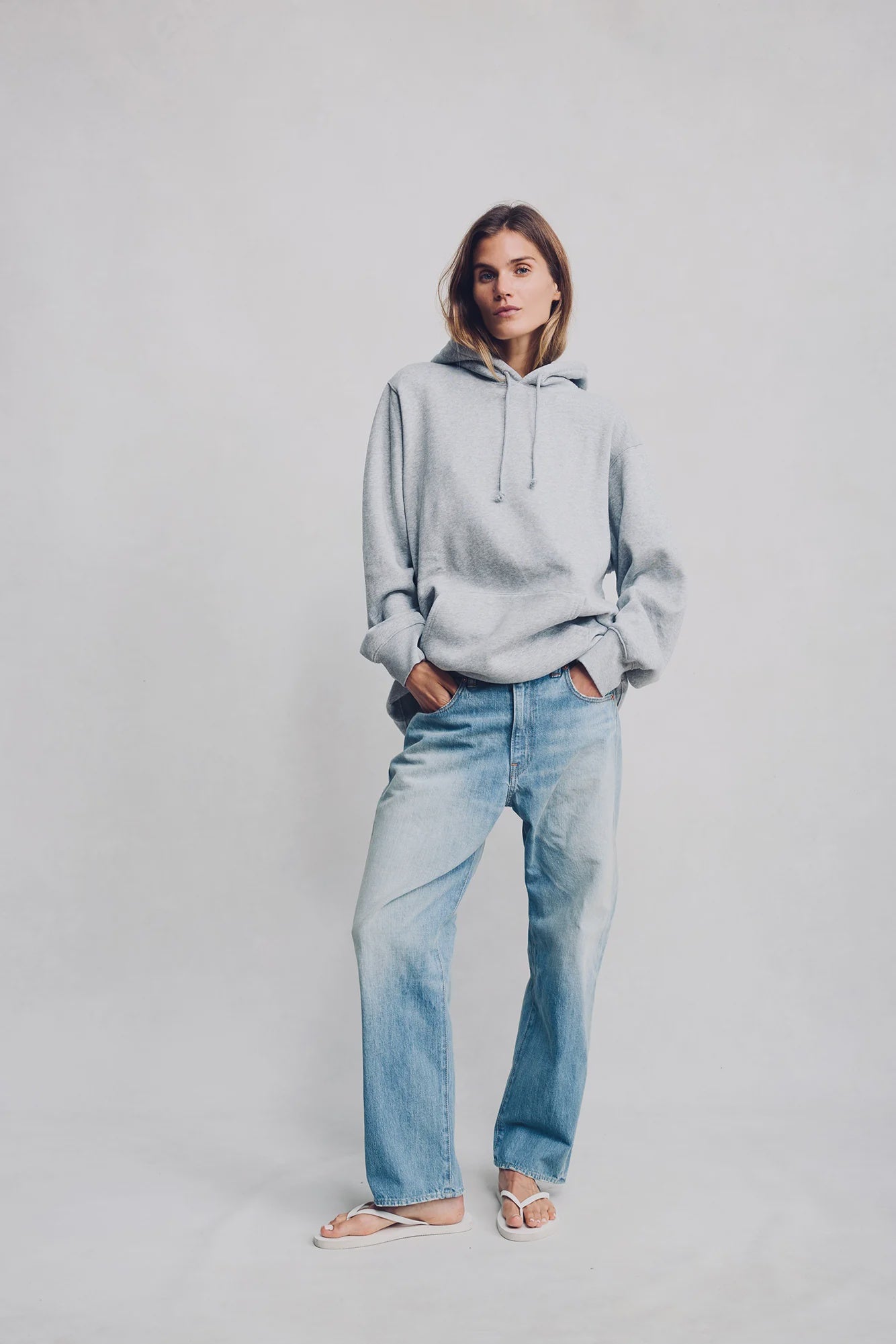 A person stands against a light background wearing a loose grey hoodie, Denimist LUCY BF JEAN MAIRE BLUE boyfriend jeans, and white sandals, hands in pockets and looking relaxed.