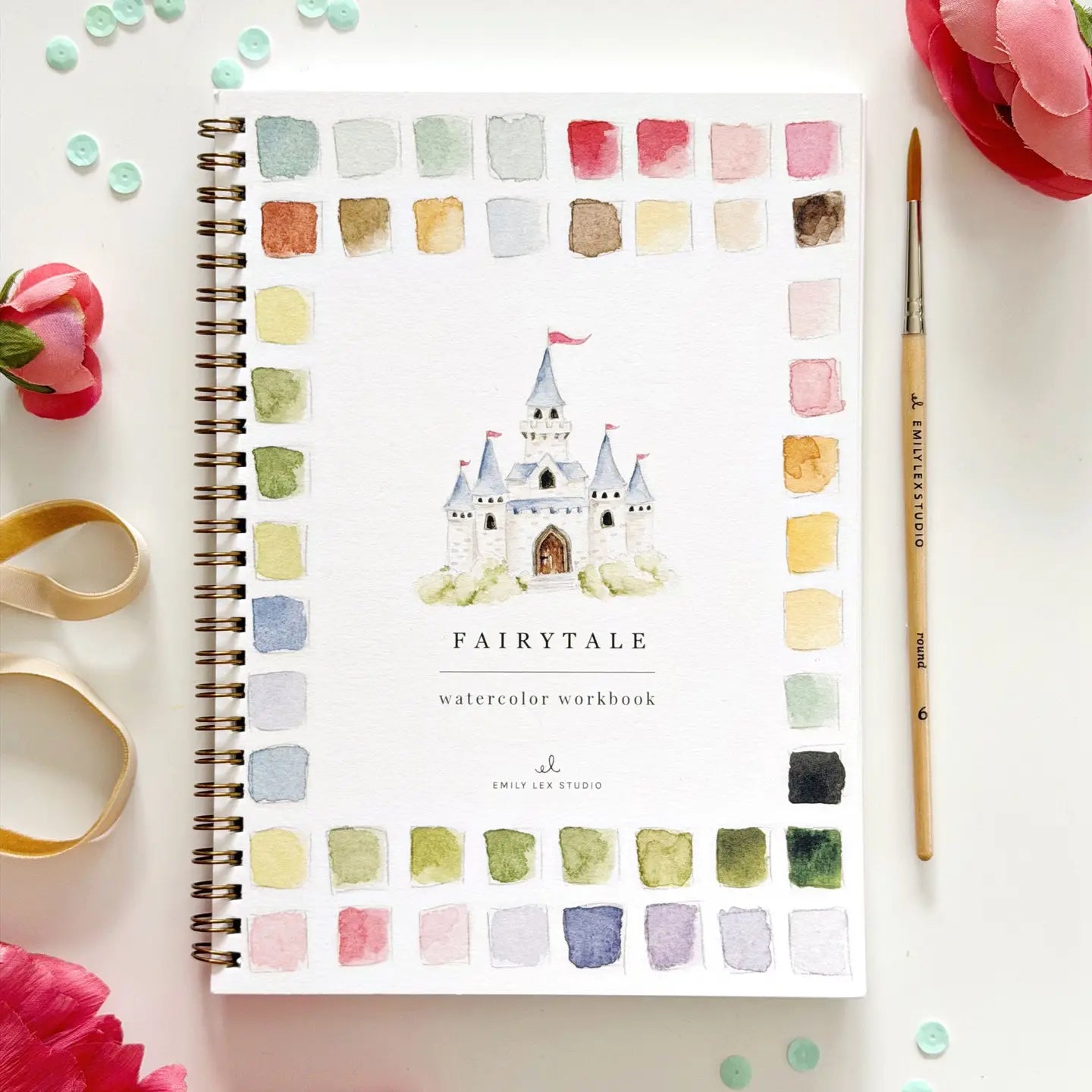 The Faire Watercolor Workbook is a spiral-bound beginner-friendly book with a castle cover and vibrant paint swatches, styled with a paintbrush, pink flowers, ribbon, and green beads on a white surface.