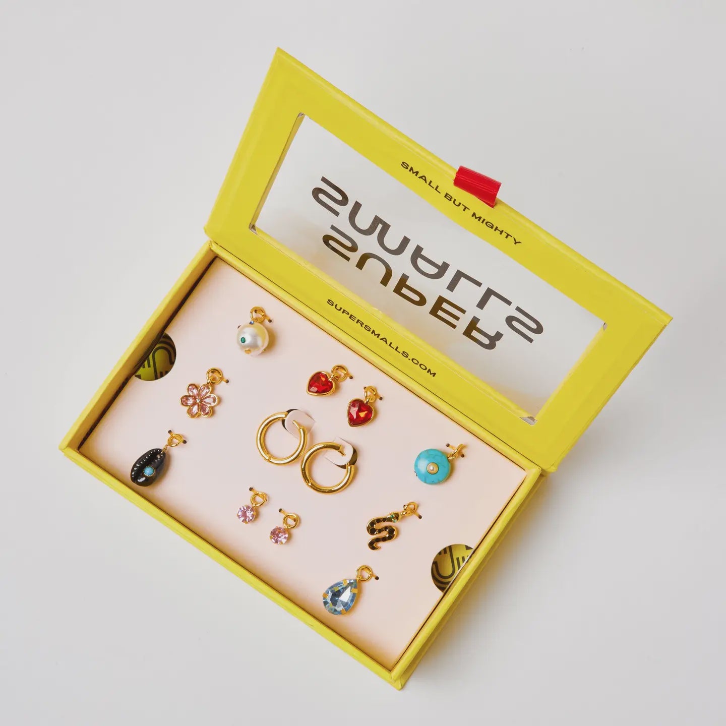 The Mix & Match Charmed Earring Set by Faire features hypoallergenic, 14K gold-plated earrings in heart, flower, and snake shapes, arranged in a yellow jewelry box labeled "Small but Mighty" with a pink interior.