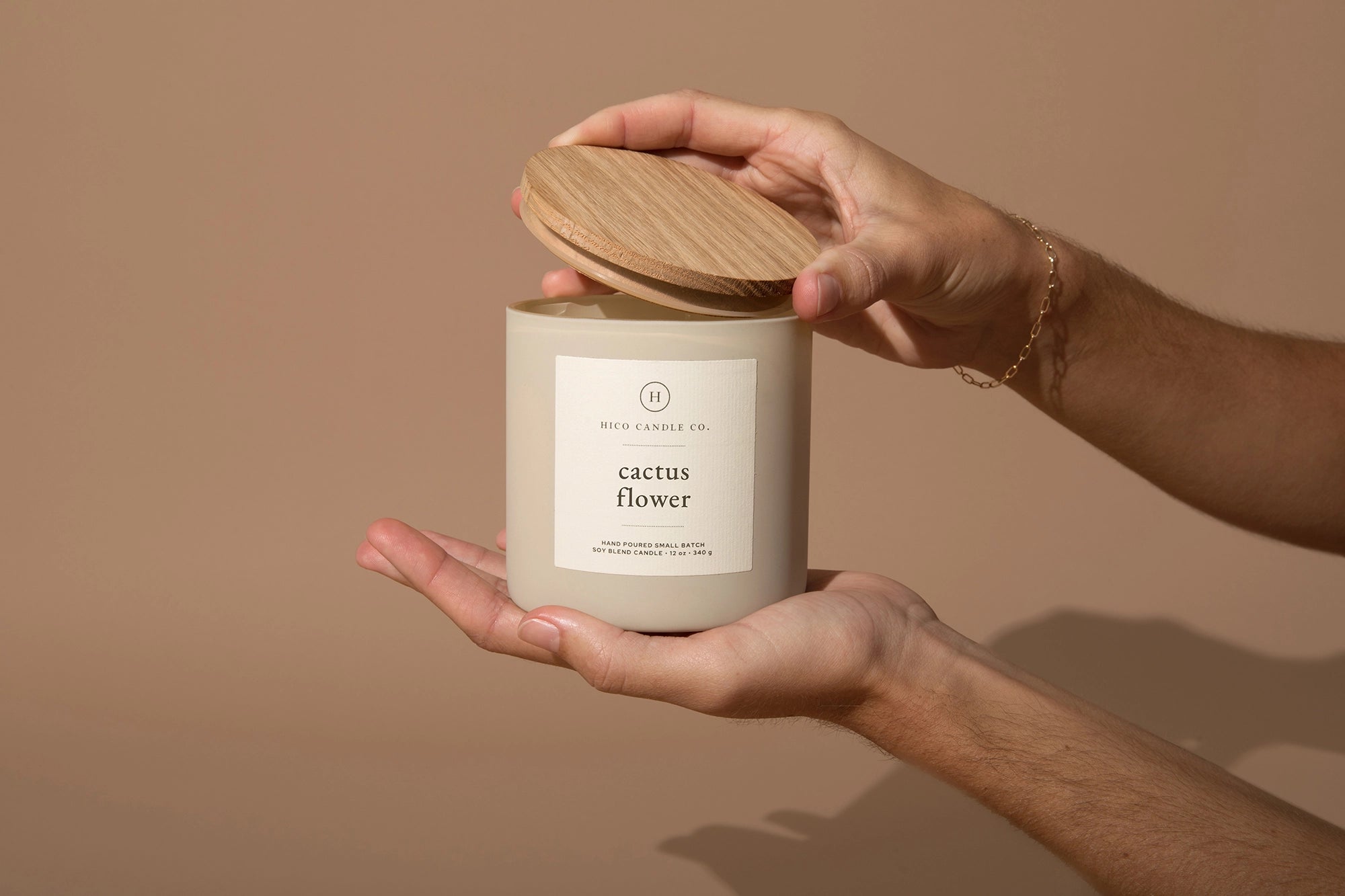 A person holds the Faire "Cactus Flower - 14oz. Candle" in one hand and lifts its wooden lid with the other, against a plain light brown background.