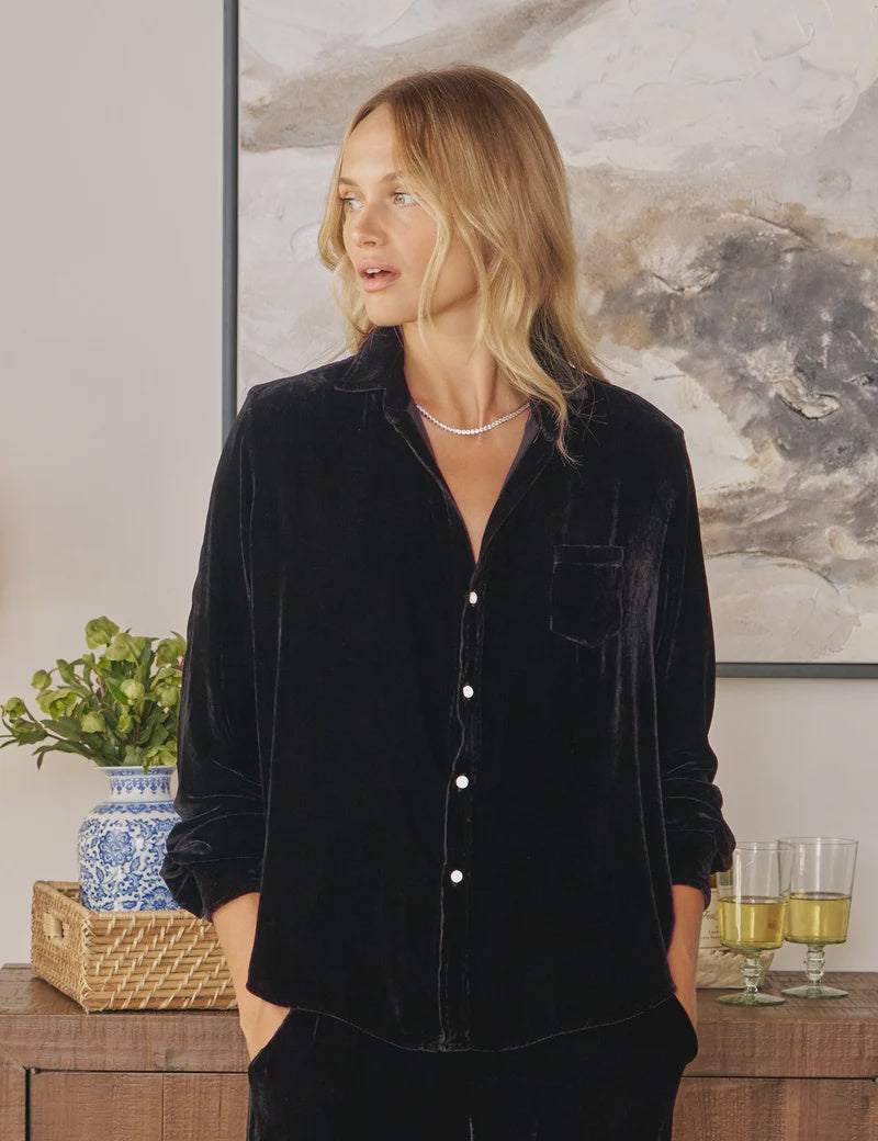 A blonde woman wears the Tuscany Getaway Eileen shirt by Frank & Eileen—an Italian washed velvet button-up—standing near a wooden table with a potted plant, glasses, and a blue-and-white vase before an abstract painting.