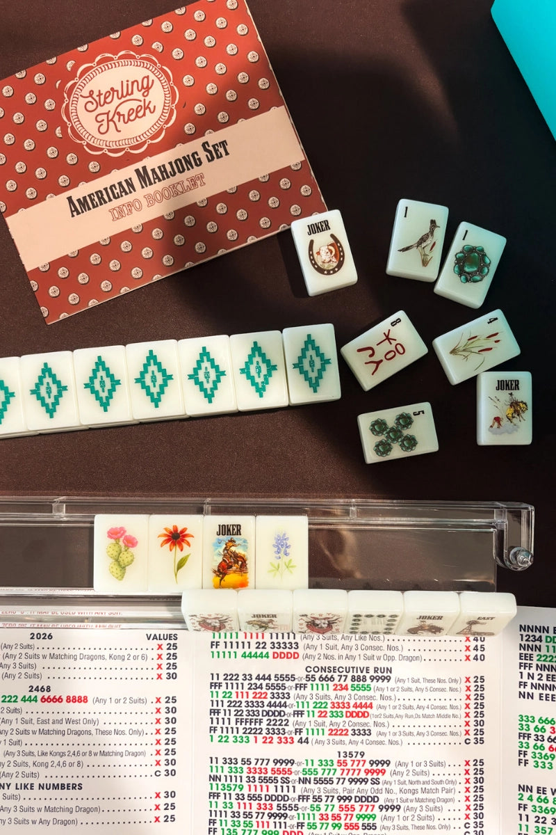 Faire Wild West Mahjong SET, featuring tiles (with jokers and character tiles), 4 racks, a bag, and a mat displayed on a brown surface with the set box, rules card, and arranged rows of tiles visible.