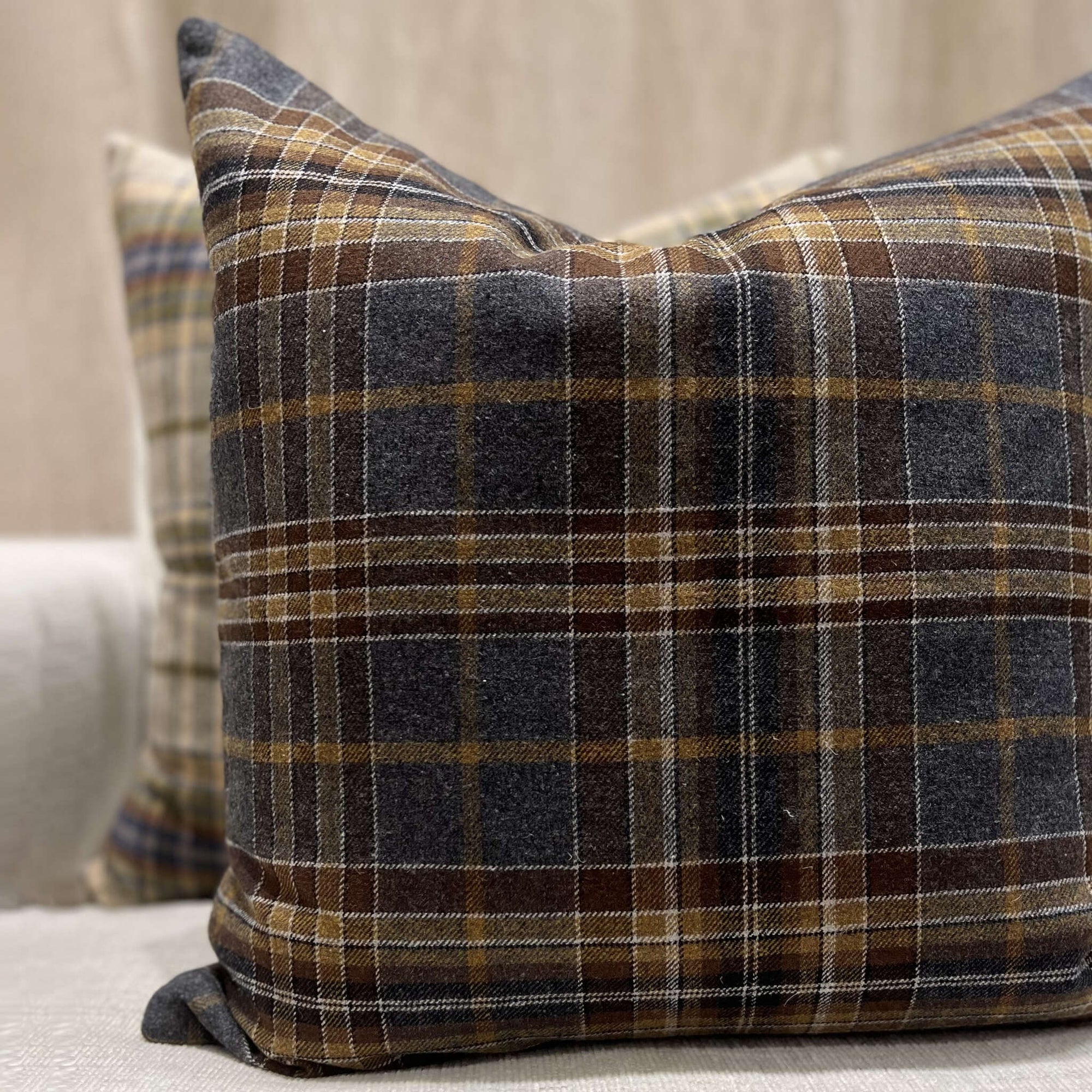 Two TL At Home Durrell Pillows 24X24, featuring brown, gray, and white plaid wool stripes, rest on a light-colored sofa. The front pillow is in focus; the one behind is slightly blurred.