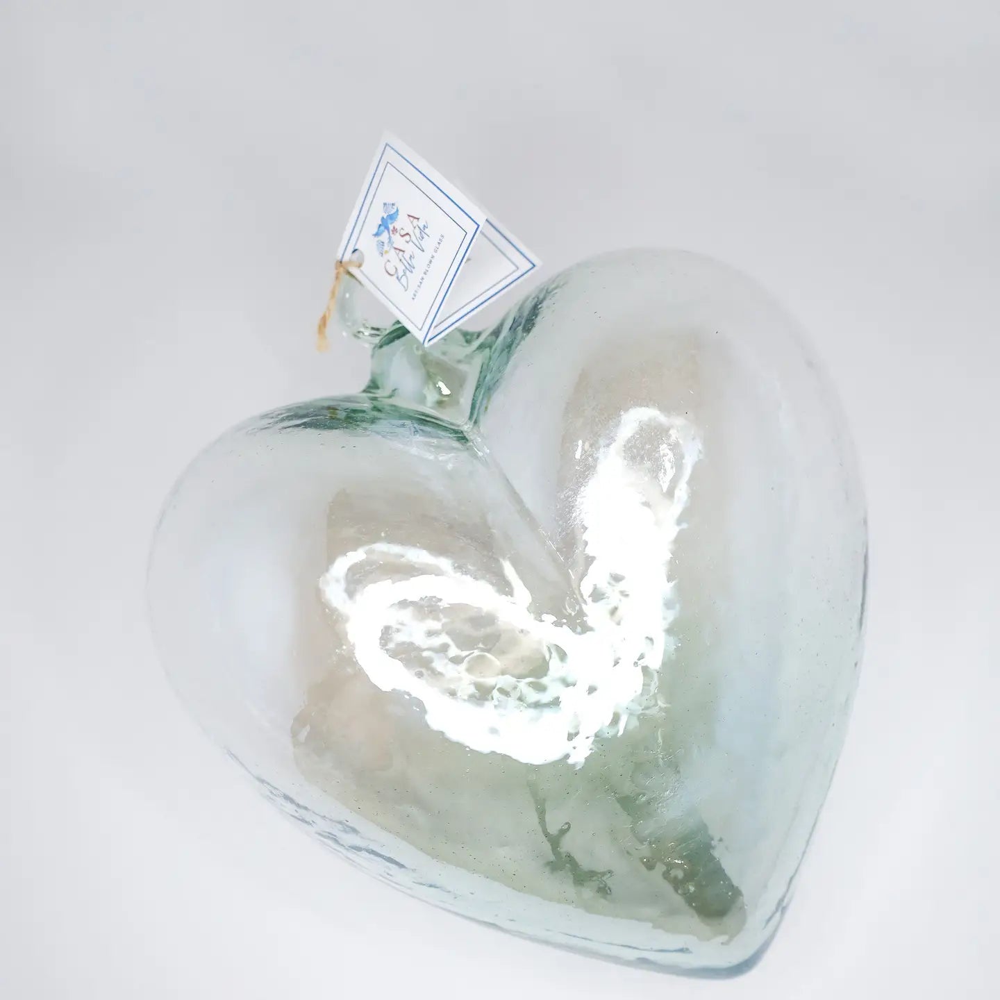 The Glass Heart Small by Faire is a clear, handcrafted blown glass heart-shaped bottle with a cork stopper, resting on a white surface. A small tag adorns its neck, while the reflective sheen and white highlights beautifully capture the finesse of Mexican tradition.