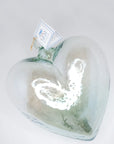 The Glass Heart Small by Faire is a clear, handcrafted blown glass heart-shaped bottle with a cork stopper, resting on a white surface. A small tag adorns its neck, while the reflective sheen and white highlights beautifully capture the finesse of Mexican tradition.