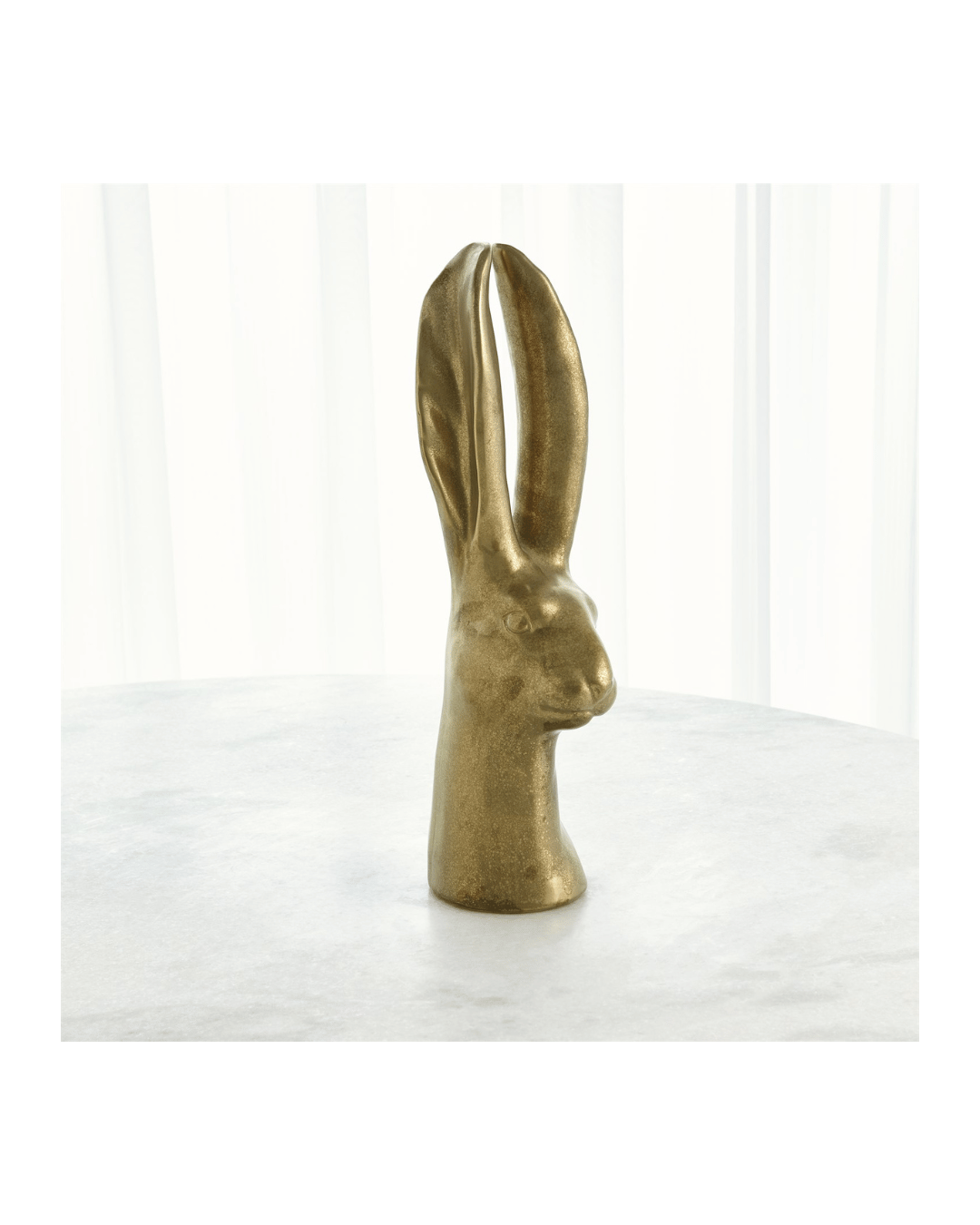 The Rabbit-Matte Gold Large sculpture by Global Views, with its elongated ears and matte gold finish reminiscent of 19th-century art, sits elegantly on a white marble surface. Softly lit sheer white curtains in the background enhance this captivating conversational piece.