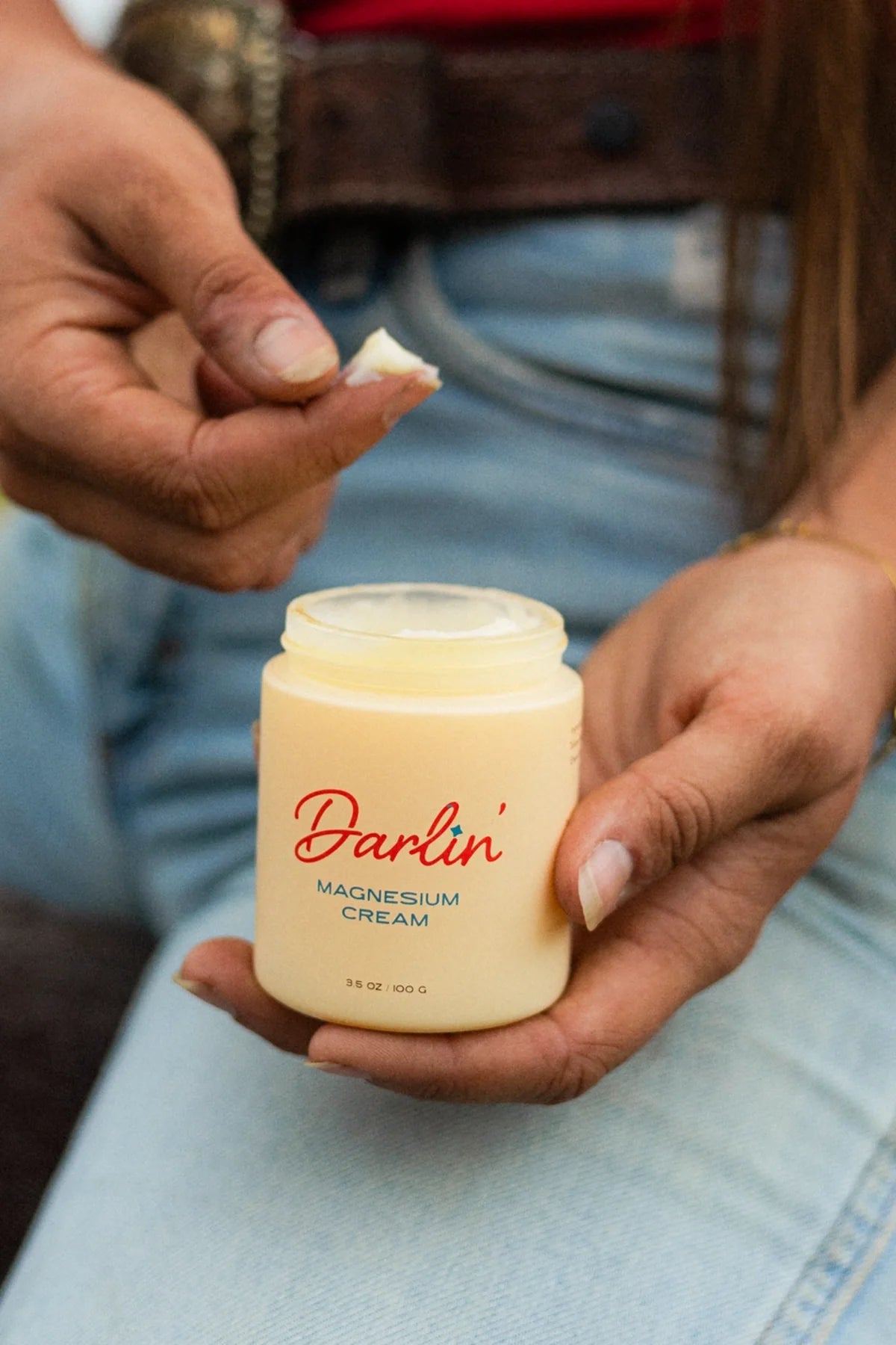 A person in blue jeans and a belt scoops Not specified Magnesium Cream from a jar, dipping their hand into the rich blend of essential oils and beef tallow.