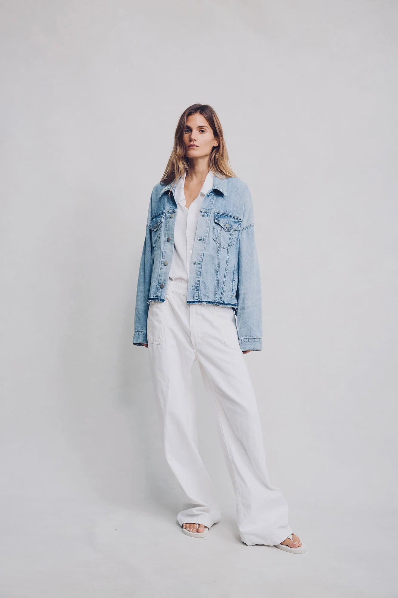 A woman with straight, shoulder-length hair wears the Denimist OVERSIZED TRUCKER SHACKET—a 100% cotton, light stone washed denim piece—over a white shirt and loose white pants with open-toed sandals, standing against a light background.