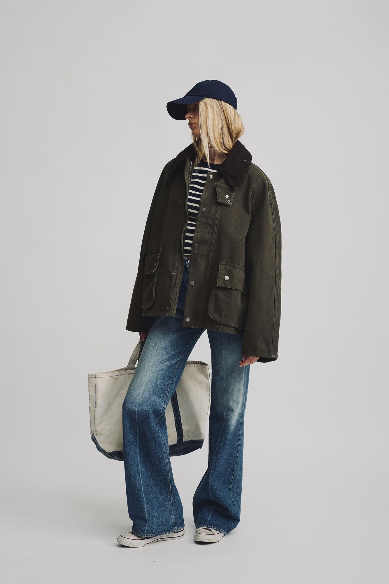 A person stands against a plain background wearing Denimist's O/S Field Jacket, a navy cap, striped shirt, flared jeans, sneakers, and holding a large white tote bag.