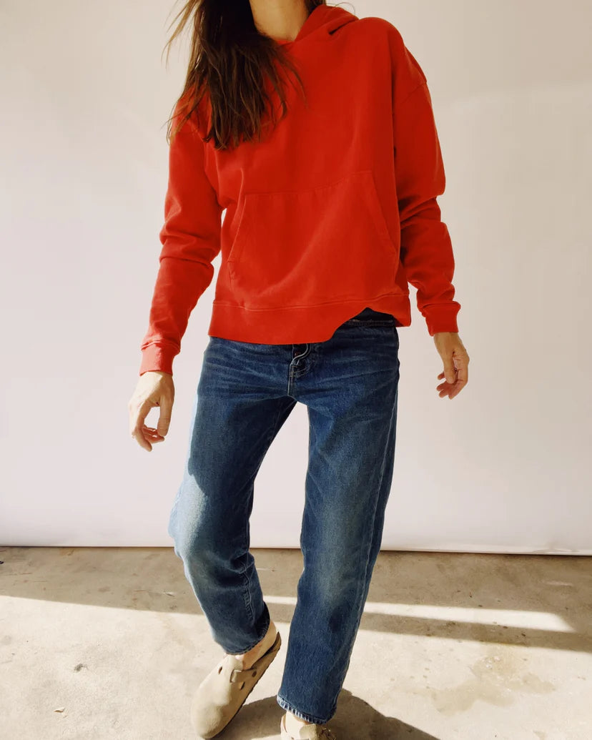 A person dressed in a red &quot;The Hoodie&quot; from Hey Gang, designed for a unisex fit, paired with blue jeans and beige shoes, stands against a plain background. The 100% cotton hoodie drapes comfortably as their hands rest casually at their sides, obscuring their face from view.