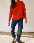 A person dressed in a red "The Hoodie" from Hey Gang, designed for a unisex fit, paired with blue jeans and beige shoes, stands against a plain background. The 100% cotton hoodie drapes comfortably as their hands rest casually at their sides, obscuring their face from view.