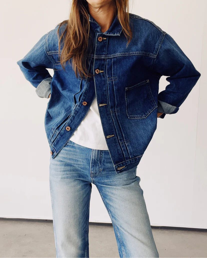 A person exudes confidence with hands on hips, wearing Hey Gang's "The Denim Jacket" along with jeans, standing against a plain white background.