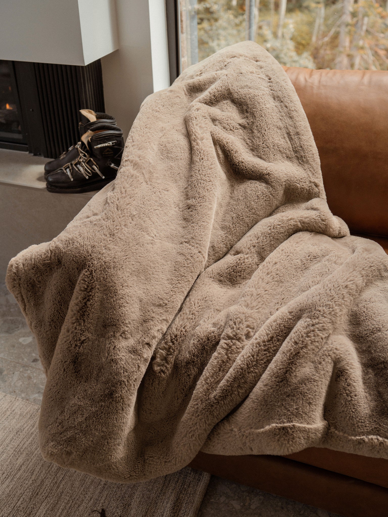 A person is wrapped in a beige Cozy Earth Cuddle Blanket with ultra-plush pile on a brown leather couch near a window, while black boots rest nearby.