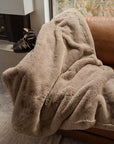 A person is wrapped in a beige Cozy Earth Cuddle Blanket with ultra-plush pile on a brown leather couch near a window, while black boots rest nearby.