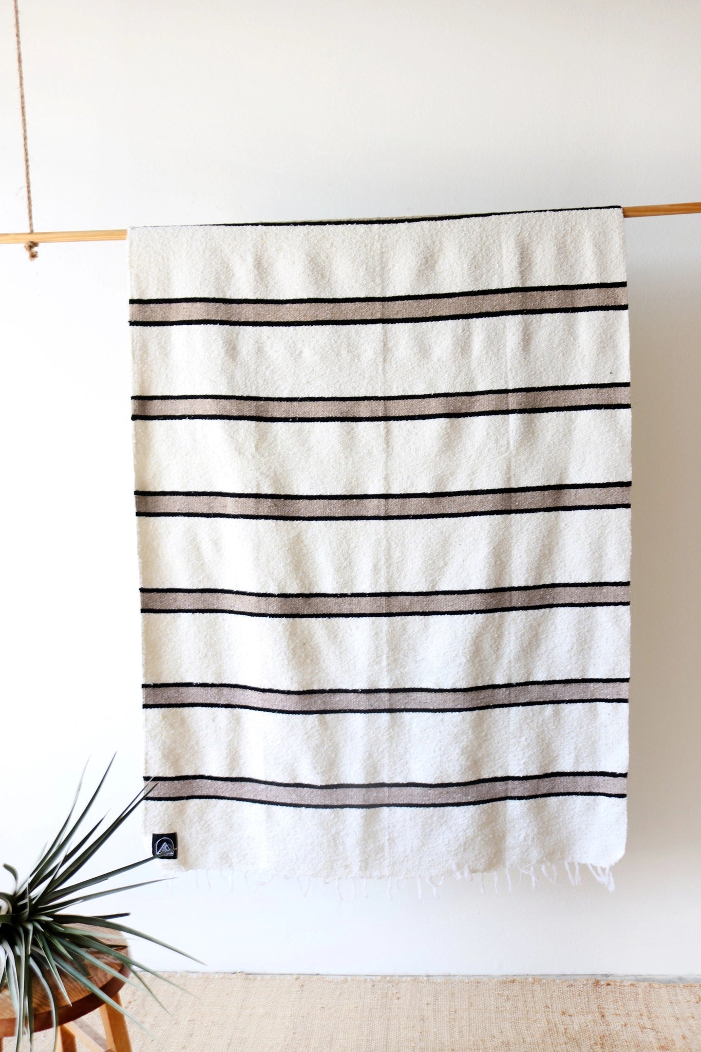The Faire TT Handwoven Blanket, featuring eco-friendly white fabric with black horizontal stripes, hangs on a wooden rod against a plain wall. A green plant appears in the corner and a wooden stool is partially visible.