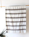 The Faire TT Handwoven Blanket, featuring eco-friendly white fabric with black horizontal stripes, hangs on a wooden rod against a plain wall. A green plant appears in the corner and a wooden stool is partially visible.