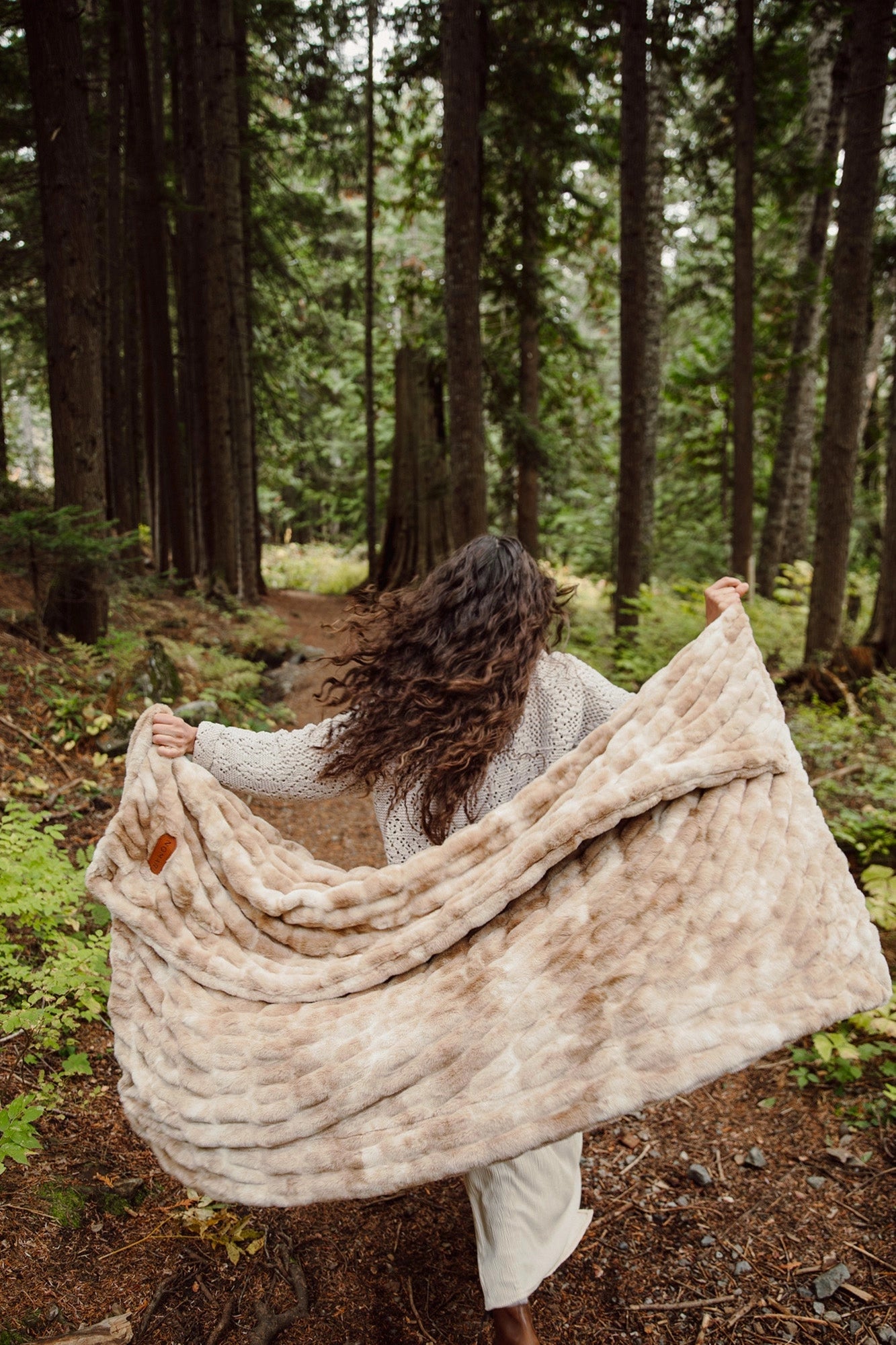 A person with long, wavy hair strolls through a forest, surrounded by tall trees and lush greenery, wrapped in the plush Cascade Throw Blanket from Faire.
