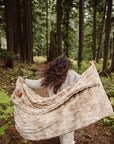 A person with long, wavy hair strolls through a forest, surrounded by tall trees and lush greenery, wrapped in the plush Cascade Throw Blanket from Faire.
