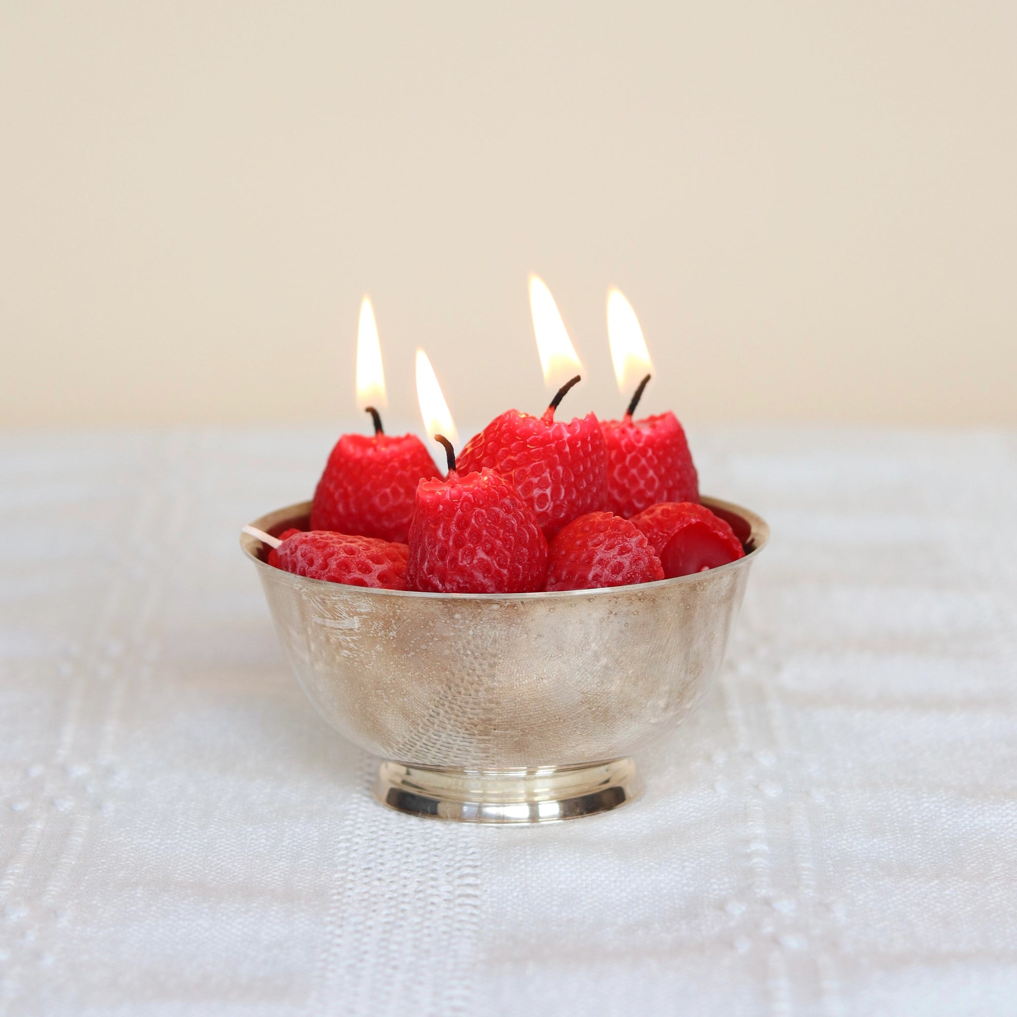 A silver bowl holds several Faire Strawberry Birthday Candles, each shaped like a red strawberry and made of beeswax with lit wicks, displayed on a white textured surface.