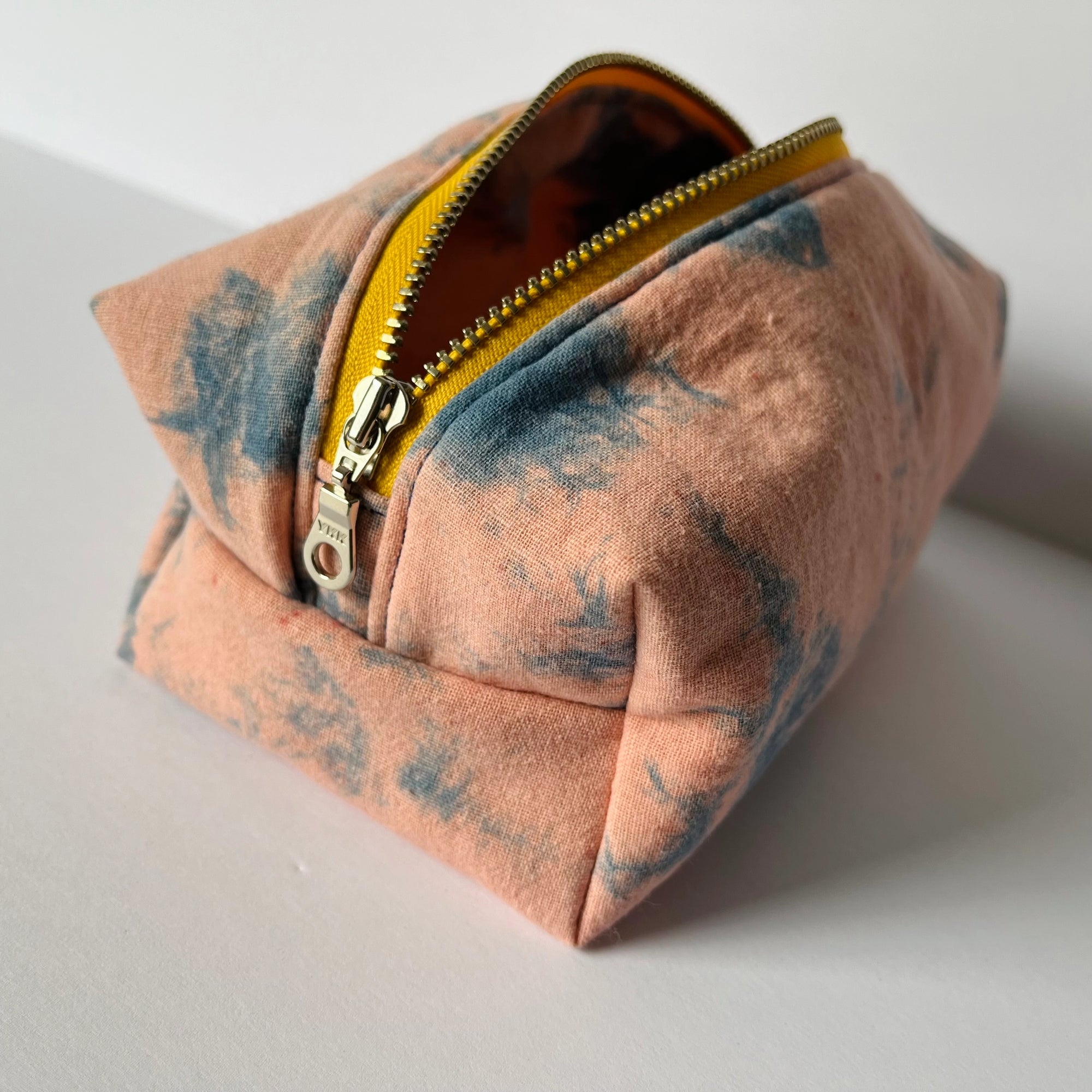 The Faire Dyed Toiletries Bag is a small, square pouch with a yellow zipper, crafted from hand-dyed cotton in soft peach and blue-gray tie-dye—perfect for desk organization on any white surface.