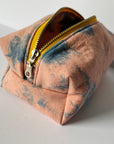 The Faire Dyed Toiletries Bag is a small, square pouch with a yellow zipper, crafted from hand-dyed cotton in soft peach and blue-gray tie-dye—perfect for desk organization on any white surface.