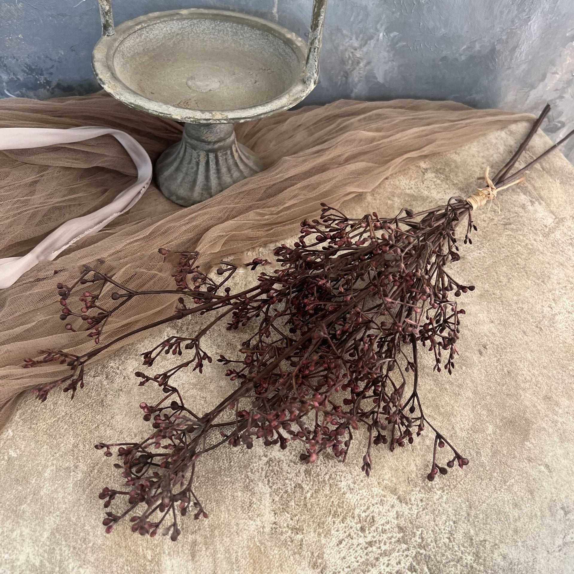 The Faire 22'' Faux Seeded Baby's Breath Bunch, tied with twine, is displayed on a textured beige surface next to a vintage metal pedestal dish and sheer brown fabric.