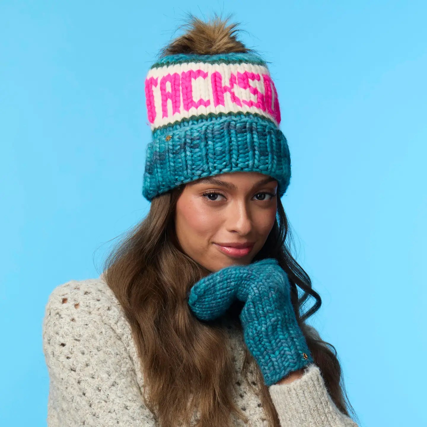 A woman wearing the Faire Knit Ski Hat—blue merino wool with faux fur pom pom and bold pink lettering—plus matching mittens and a cream sweater, poses against a blue background.