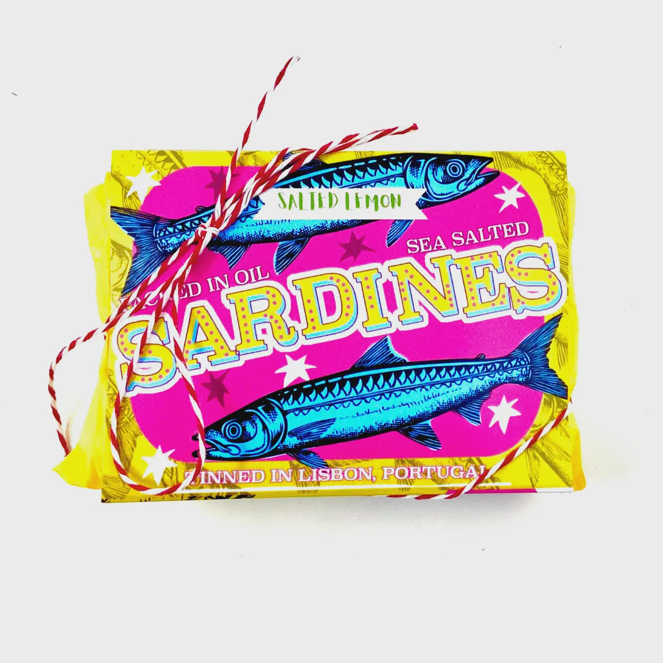 The Sardine Tin Luxury Soap by Faire is wrapped in red and white twine. The vibrant label, featuring blue fish illustrations, reads "Salted Lemon Sea Salted Sardines" with "Tinned in Lisbon, Portugal." Its bright yellow and magenta design resembles luxury soap bars.