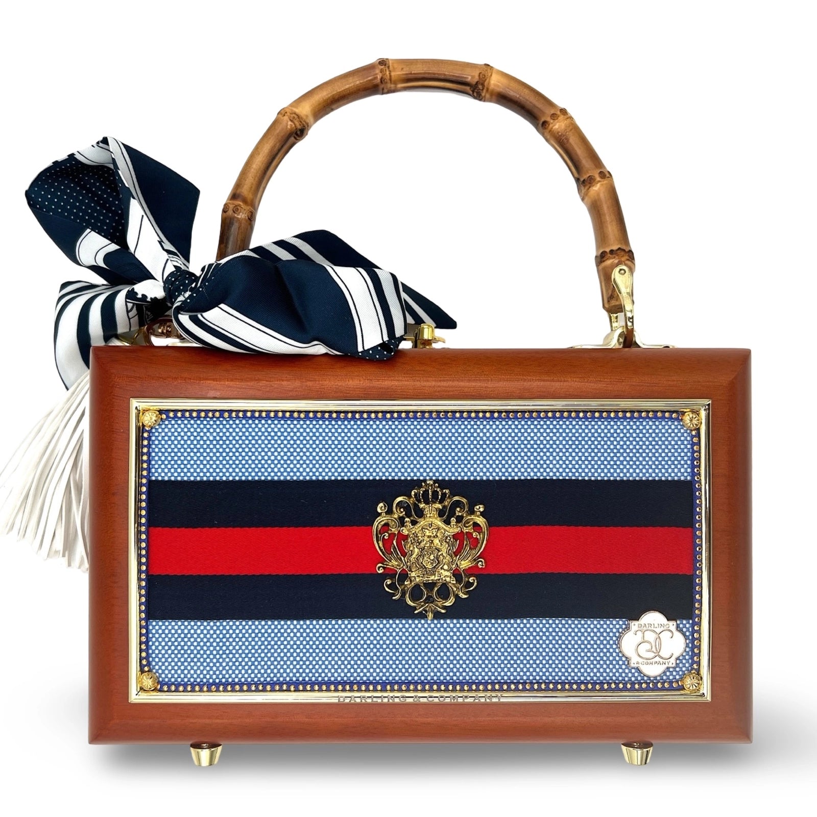 The Faire Grand Old Flag Bag is a rectangular, hand-finished designer piece with a bamboo handle, gold crest, navy/black/red striped panel, and a black-and-white striped scarf with a white tassel tied to the handle.