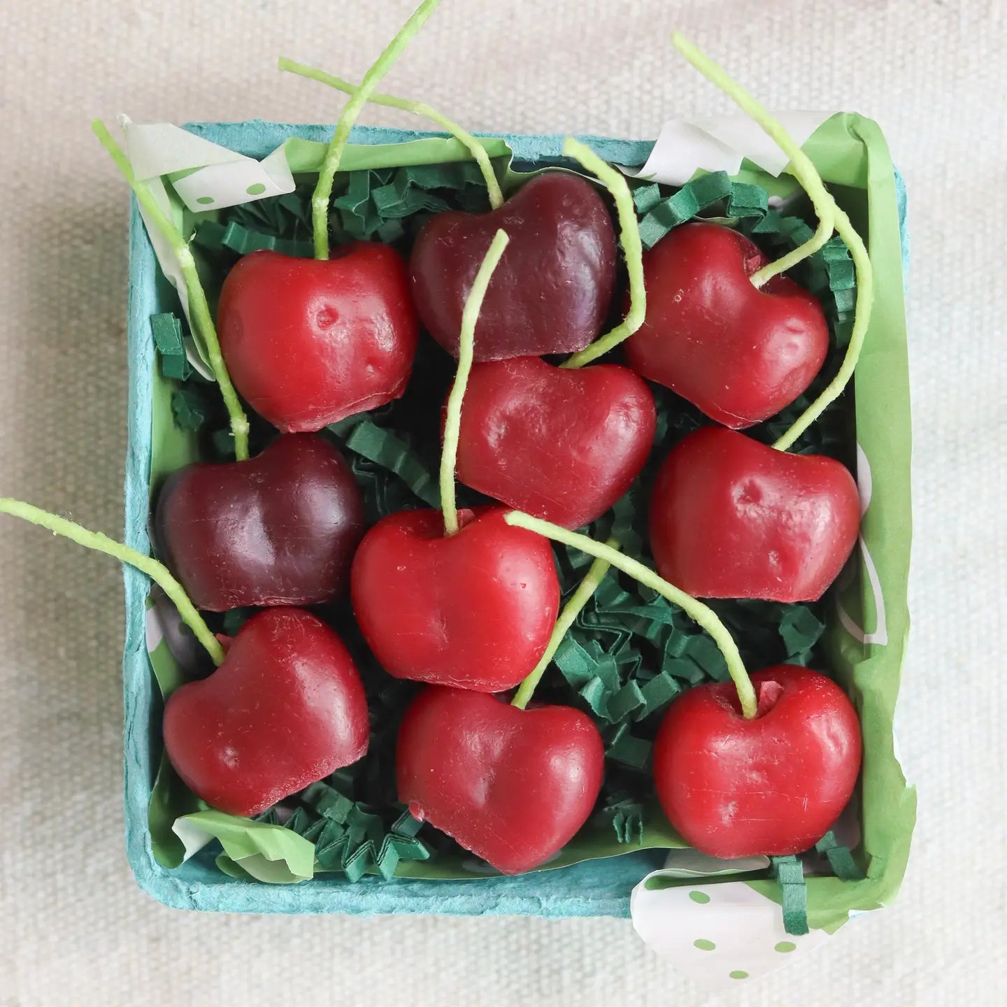 A small blue basket by Faire holds Cherry Beeswax Candles shaped like red and dark red hearts with green candy stems, arranged on green paper shreds.