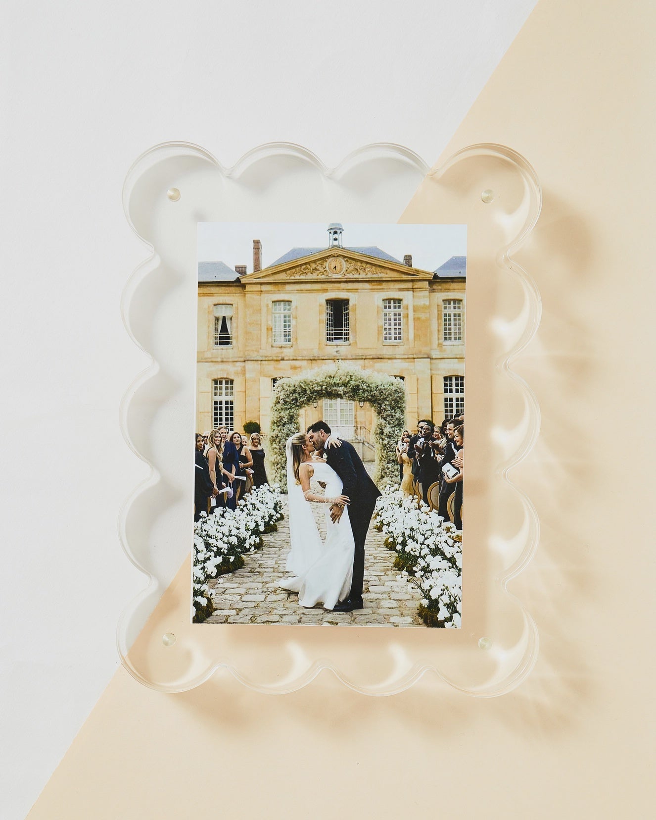 Acrylic Frame by Faire: A captivating photo of a newlywed couple kissing, embraced by guests and white flowers in front of a grand building with arched windows and stone pathway, enhanced by a decorative magnetic border for an elegant touch.