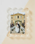 Acrylic Frame by Faire: A captivating photo of a newlywed couple kissing, embraced by guests and white flowers in front of a grand building with arched windows and stone pathway, enhanced by a decorative magnetic border for an elegant touch.