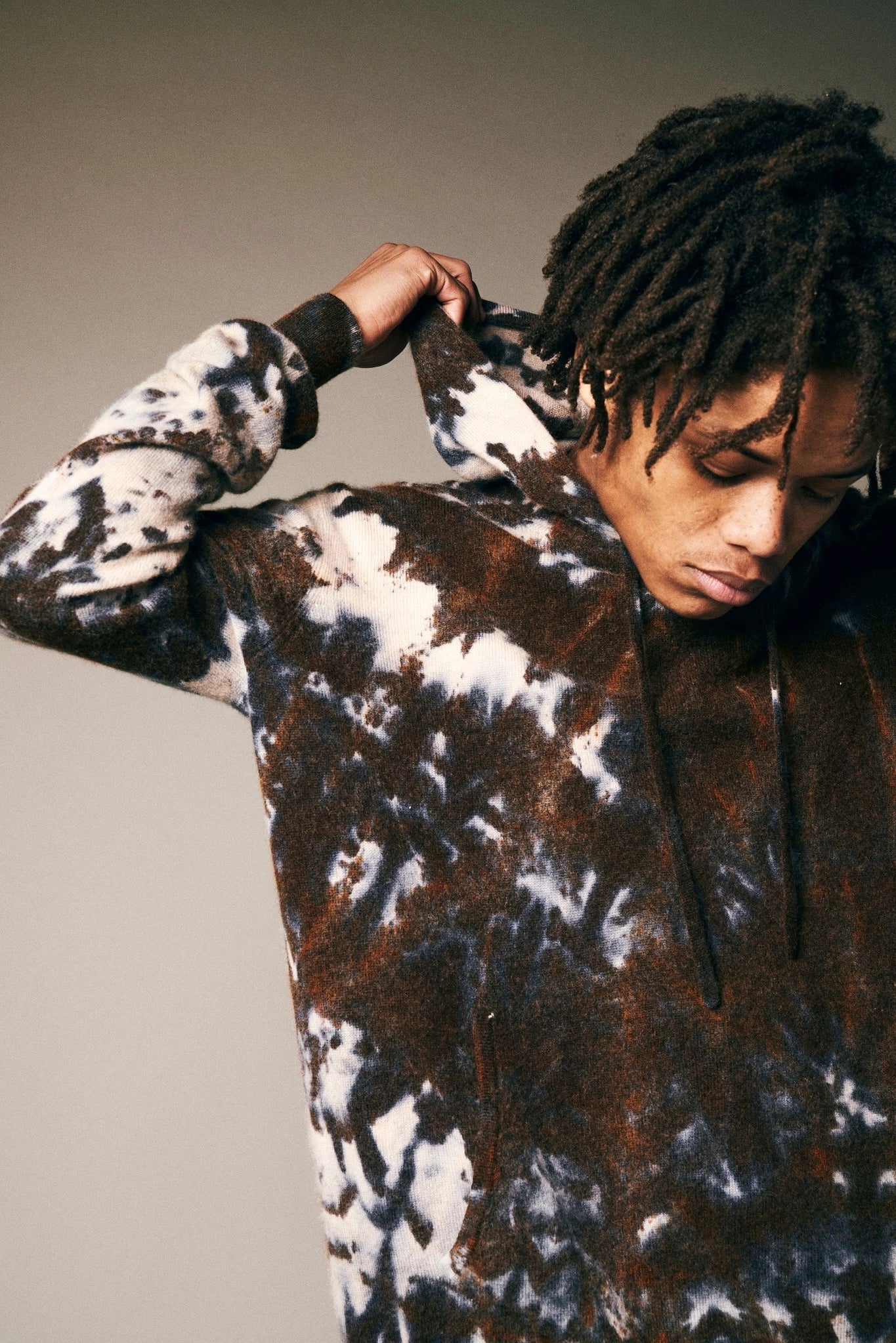 A person with medium-length dreadlocks pulls up the hood of a Riverside Tool & Dye Cashmere Hoodie in hand-painted brown, white, and black tie-dye, looking down against a neutral background.
