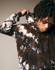 A person with medium-length dreadlocks pulls up the hood of a Riverside Tool & Dye Cashmere Hoodie in hand-painted brown, white, and black tie-dye, looking down against a neutral background.