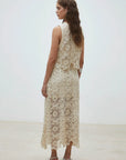 A woman with long wavy hair stands in a minimalistic room, wearing the SUNCOO Fanny Skirt—an ankle-length cream skirt with intricate floral crochet and lace details—facing slightly away to showcase its elegant design.