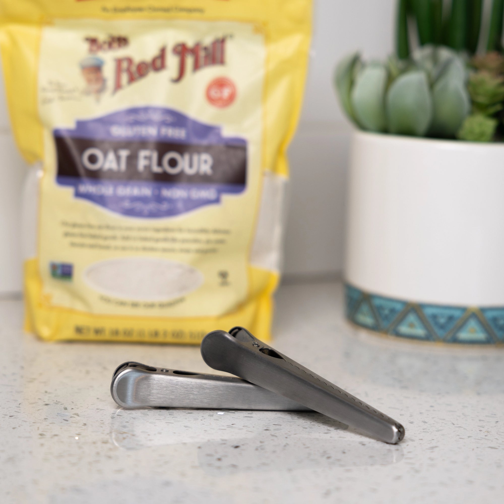 A Faire Stainless Steel Kitchen Bag Clip sits on a countertop in front of a yellow bag of Bob's Red Mill oat flour, with a potted succulent plant in the background.