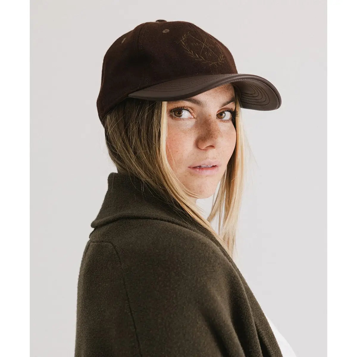 A woman with light skin and freckles models the Faire Aven Leather Ball Cap—a dark brown wool felt hat with a faux leather brim—paired with an olive sweater, glancing over her shoulder at the camera against a plain background.