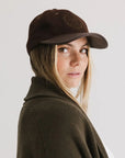 A woman with light skin and freckles models the Faire Aven Leather Ball Cap—a dark brown wool felt hat with a faux leather brim—paired with an olive sweater, glancing over her shoulder at the camera against a plain background.