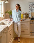 A woman in a light blue blouse and Frank & Eileen's Jameson Utility Jogger leans against a kitchen counter, holding a mug and gazing out the window. The kitchen has a marble sink and countertop, adorned with various utensils and plants.