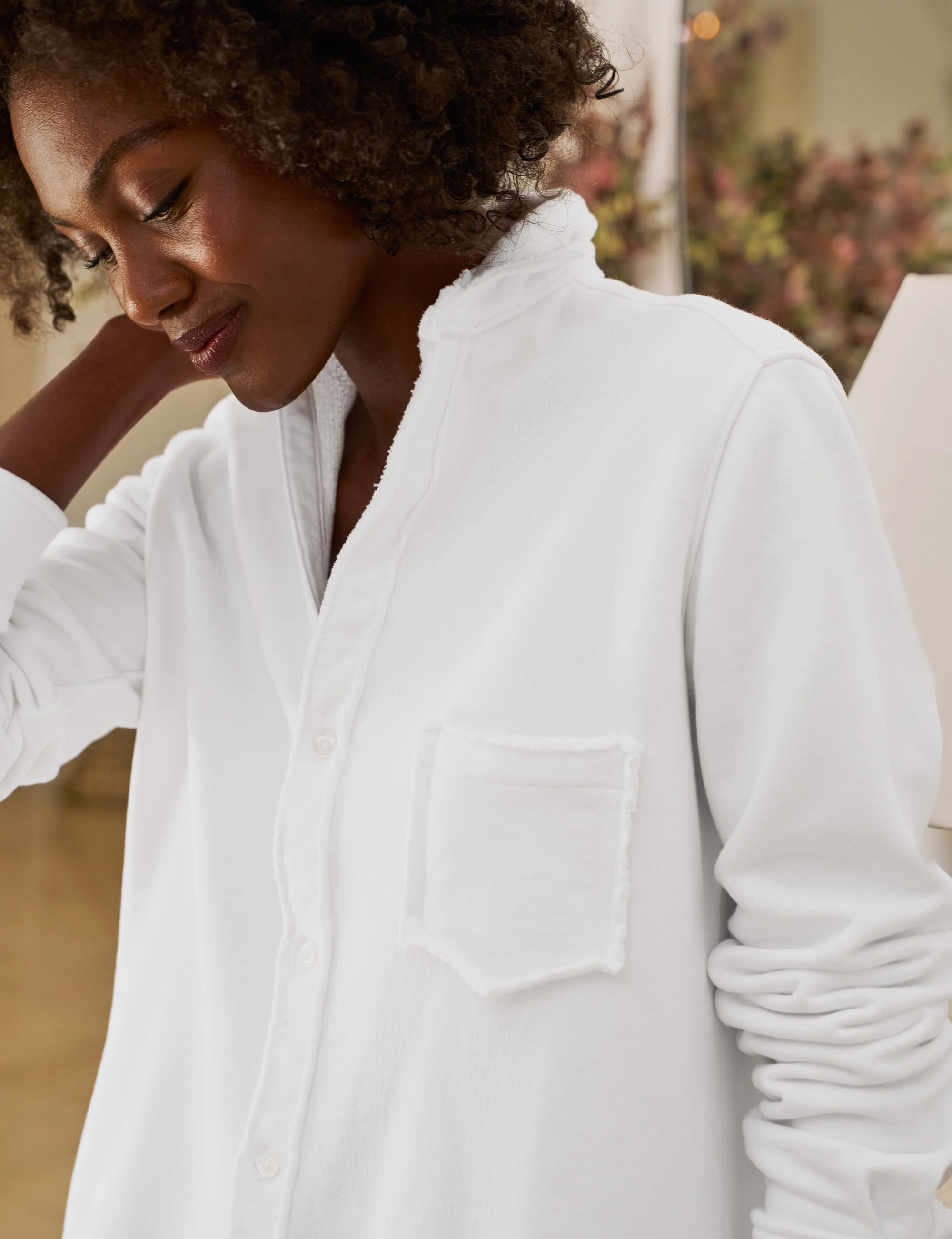 A person wearing the Frank & Eileen Eileen Sweatshirt Button Up stands indoors, gently touching the back of their head and looking down with a soft smile.