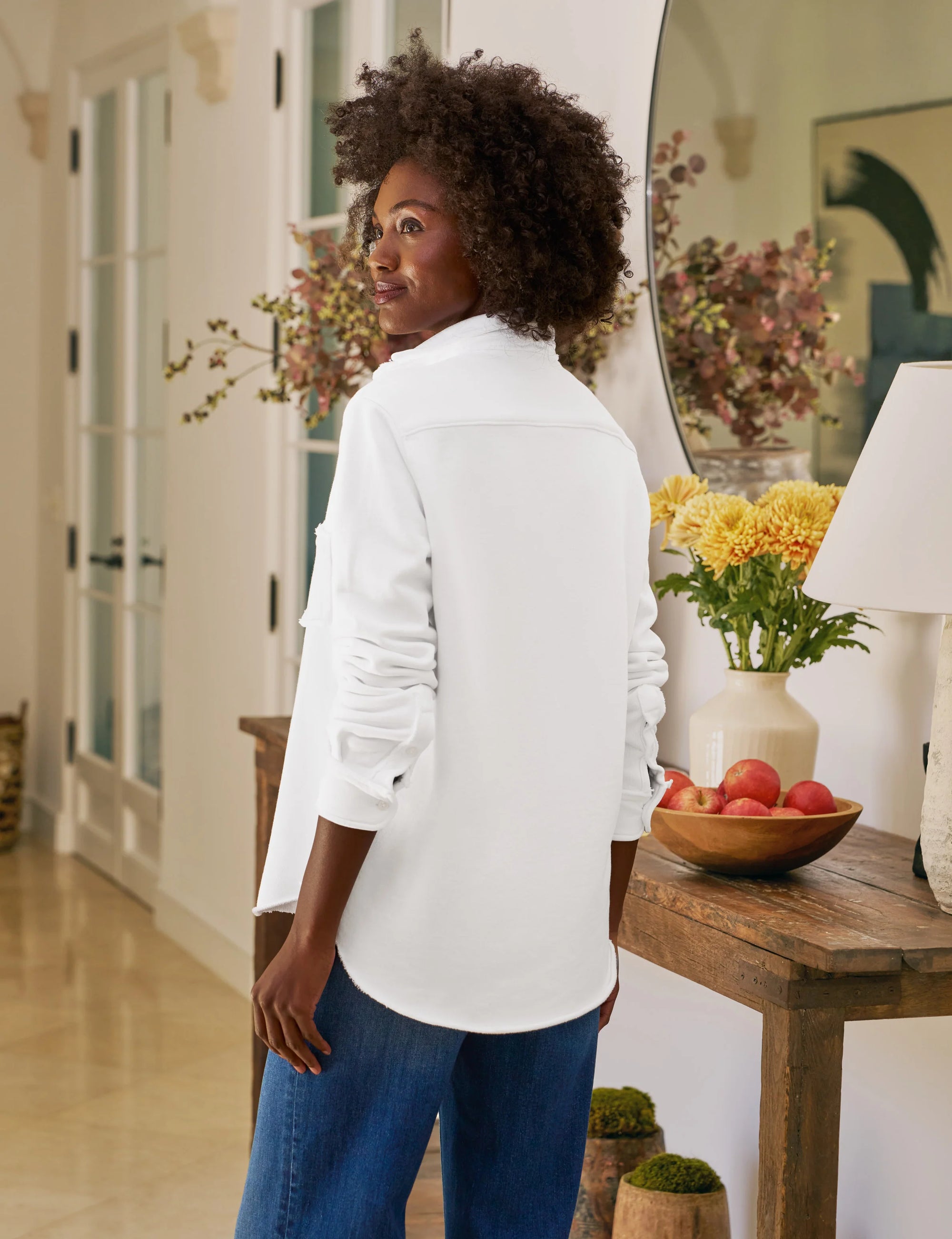 A woman with curly hair stands indoors, looking over her shoulder. She wears the Frank & Eileen Eileen Sweatshirt Button Up with blue jeans. A table with flowers, peaches, and a lamp is in the background.