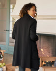 A woman with curly hair, wearing the Frank & Eileen Sweatshirt Trench Coat, stands indoors by a fireplace. She is turned slightly away from the camera and smiling. The room features cozy furniture and a light blue door with glass panels.