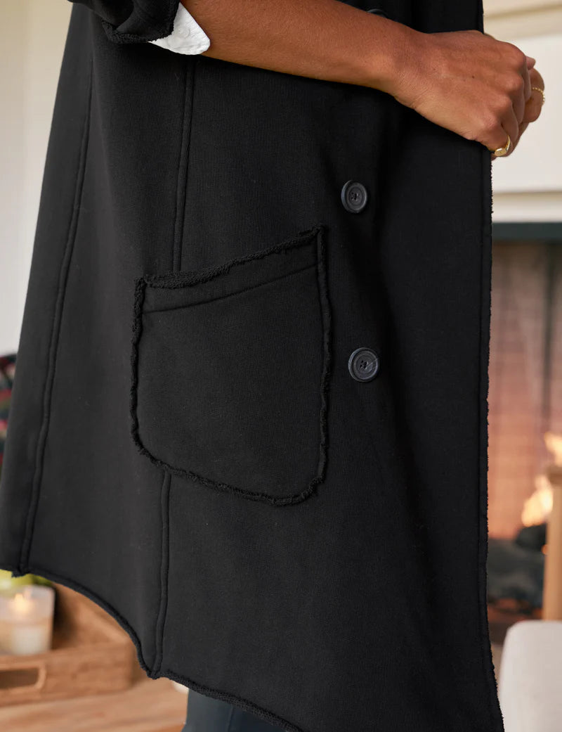 A side view of a person standing indoors by a lit fireplace, wearing the Frank & Eileen Sweatshirt Trench Coat in black with two buttons and a large front pocket; their hand rests by their side.