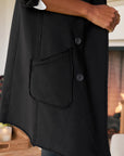 A side view of a person standing indoors by a lit fireplace, wearing the Frank & Eileen Sweatshirt Trench Coat in black with two buttons and a large front pocket; their hand rests by their side.