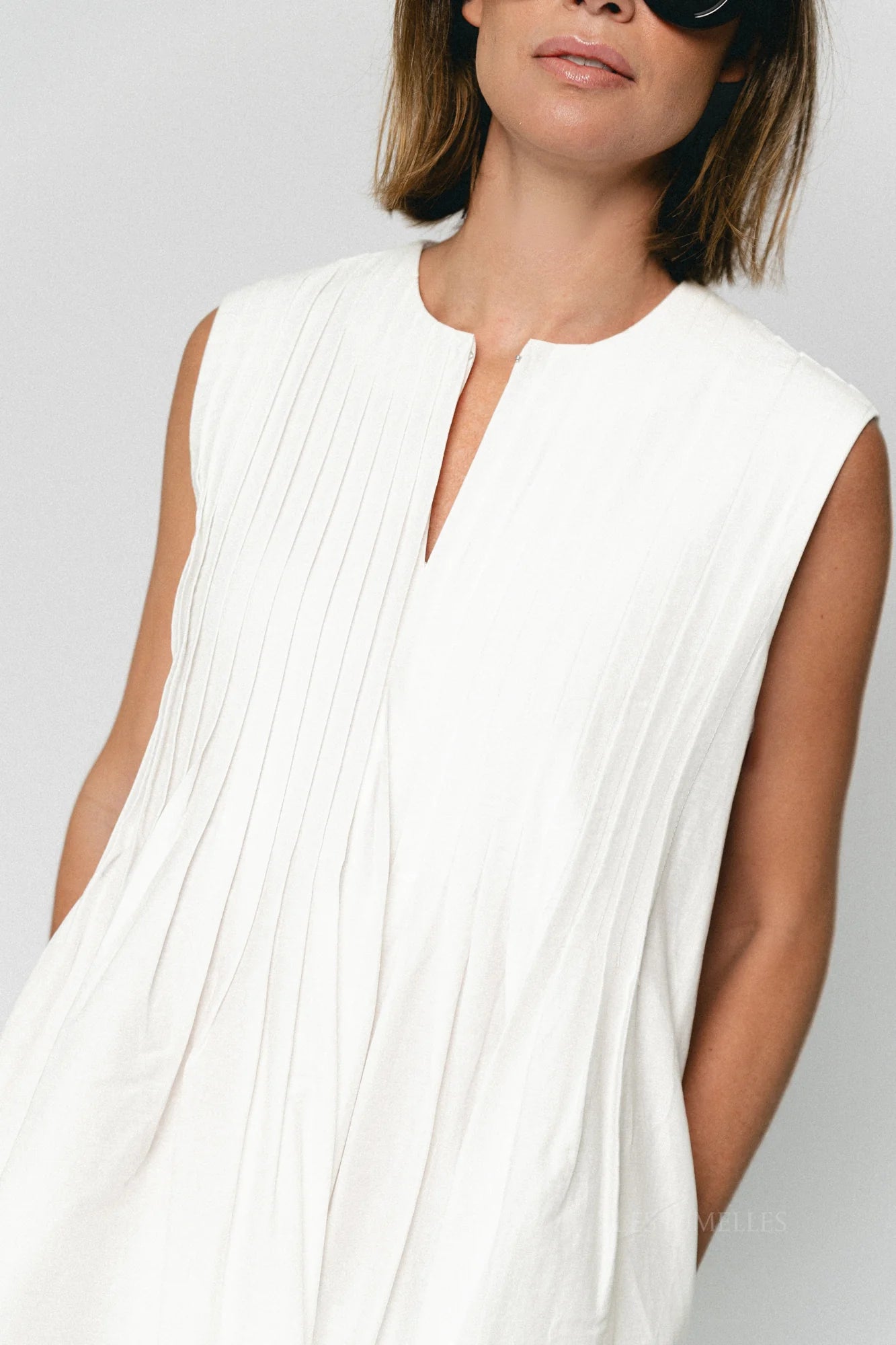 A woman wears the SUNCOO Conely Dress, a sleeveless white pleated summer dress with a relaxed fit and split neckline. She pairs it with shoulder-length brown hair and black sunglasses against a plain, light background.